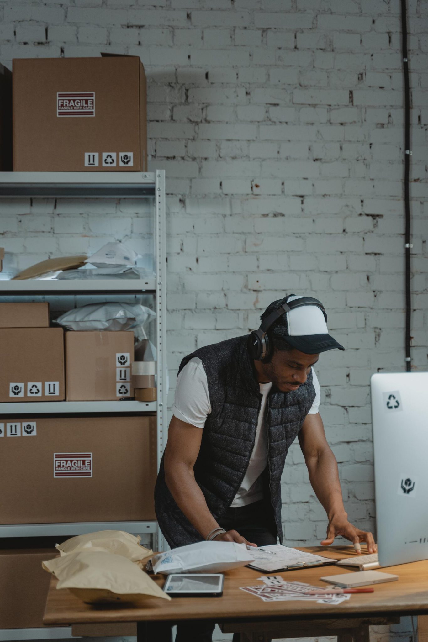 Hombre con auriculares, gorra y chaleco negro trabajando en ordenador con paquetes detrás ordenados en estantería con pared de ladrillos blanca.
